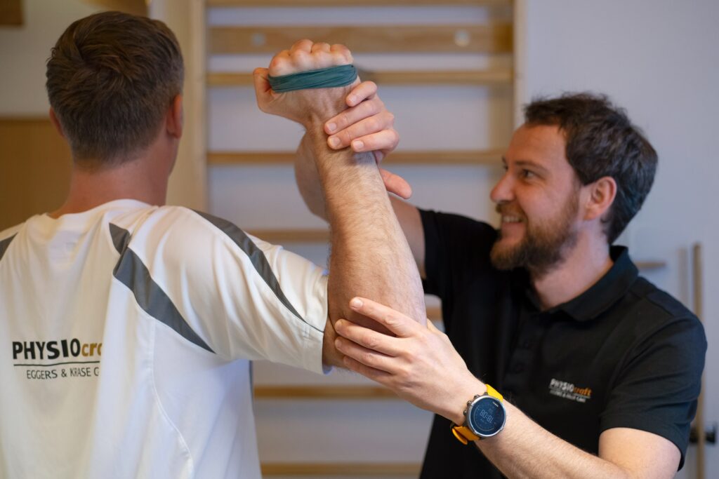 Physiotherapeut behandelt Patienten am Armgelenk, während dieser eine Übung mit seinem Arm an der Sprossenewand mit einem Terraband macht.